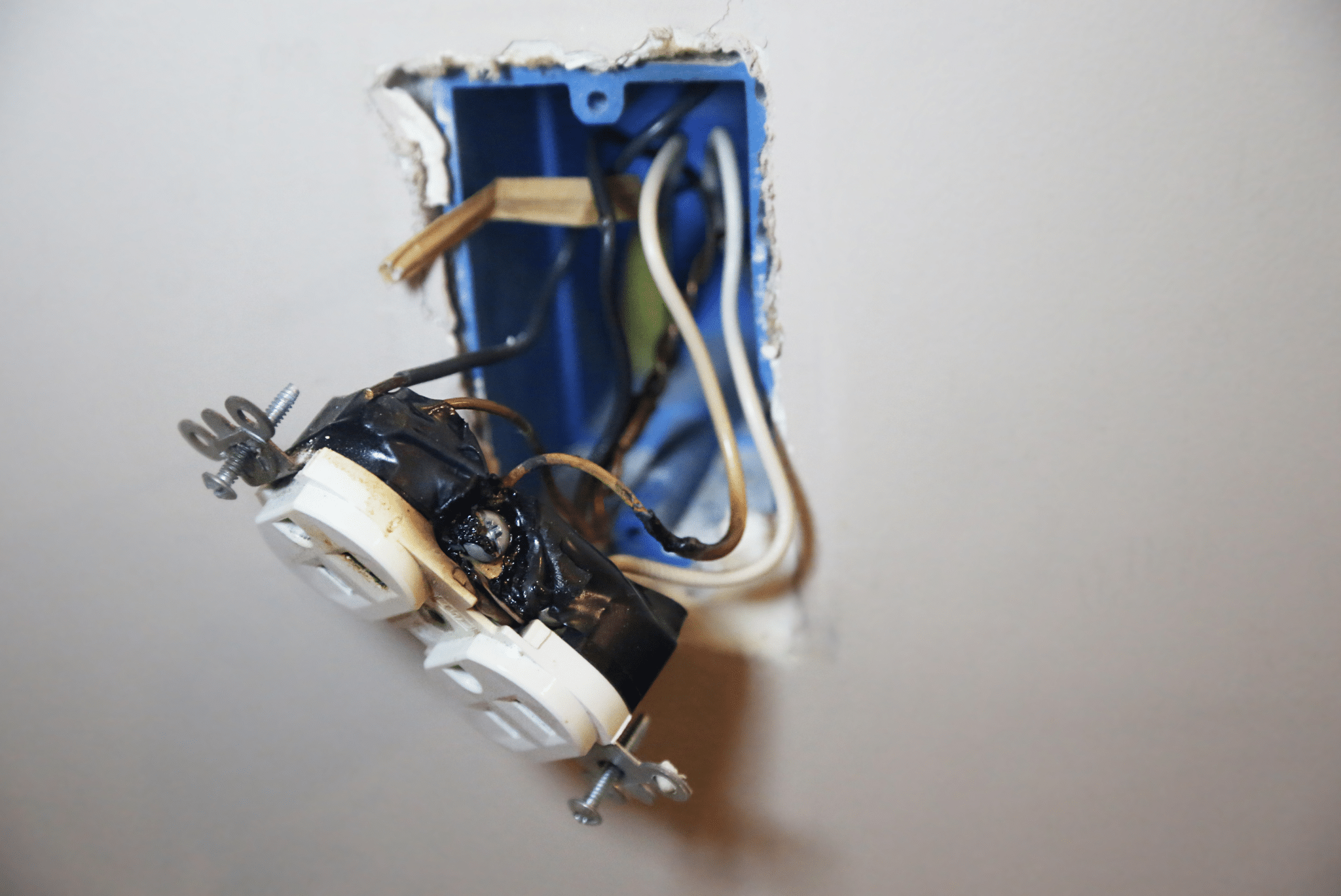 Damaged electrical outlet with exposed, burnt wires and a charred socket partially hanging from a wall. Damaged electrical outlet with exposed, burnt wires and a charred socket partially hanging from a wall.